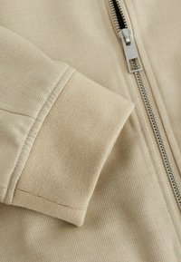 Beige lynlåsjakke med detaljer af ribbet tekstur ved mansjetten og kroppen, med en glat metallynlås og lige syninger.