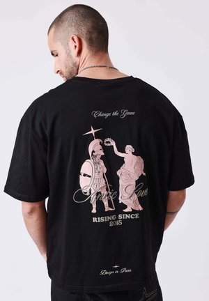 Man met geschoren hoofd draagt een zwart T-shirt met een afbeelding op de rug van twee klassieke figuren en de tekst "Change the Game" en "Rising Since 2015".