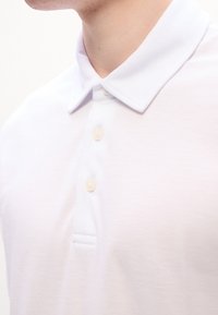 Gros plan d'une personne portant un polo blanc avec un col plié et trois boutons clairs.