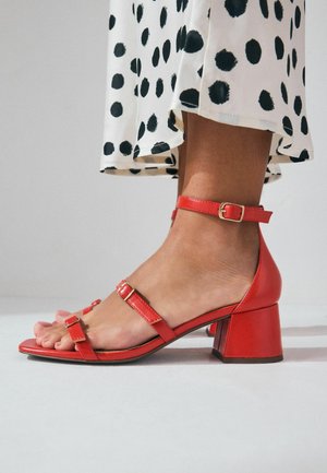 Sandalias de tacón bloque rojas de cuero con tira en el tobillo y detalles de hebilla. Diseño con tiras y punta abierta, con una textura suave, combinadas con un vestido de lunares.