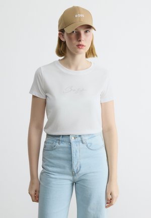 Νεαρή γυναίκα που φοράει μπεζ καπέλο με τη λέξη "BOSS", λευκό εφαρμοστό t-shirt και ανοιχτό μπλε ψηλοκάβαλο τζιν, στέκεται μπροστά σε απλό φόντο.