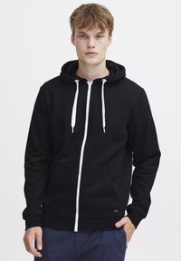 Sudadera con cremallera negra, con un bolsillo frontal, cordones blancos y una textura suave. Presenta un diseño ajustado con puños y dobladillo acanalados.