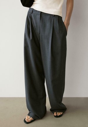 Person iført løstsiddende mørke pinstripe brede bukser med lommer og sorte flip-flops, der står på et neutralt gulv foran en ensfarvet væg.