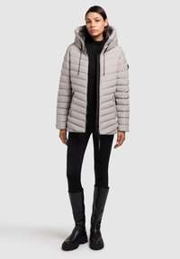 Pufferjakke i lys grå med chevron-quilting, justerbar hætte, frontlynlås og sidelommer, parret med sorte leggings og knæhøje støvler.