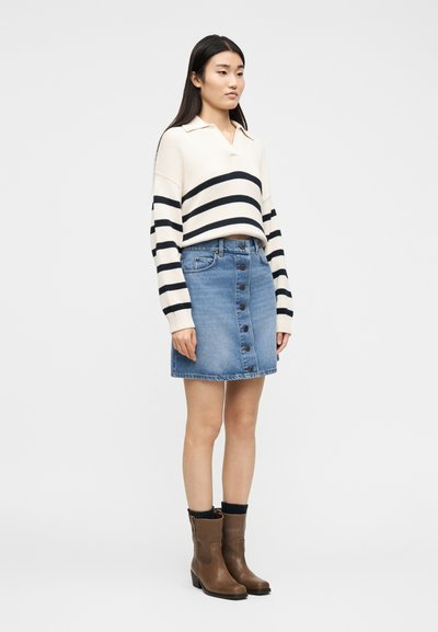 Γυναίκα που φοράε ι εκρού πουλόβερ με μαύρες ρίγες, τζιν φούστα με κουμπιά στο μπροστινό μέρος, μαύρες κάλτσες και καφέ μποτάκια αστραγάλου, στέκεται σε λευκό φόντο.