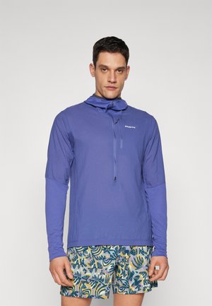 Haut léger à manches longues violet avec un col zippé et une capuche, associé à un short de bain multicolore à motifs botaniques.