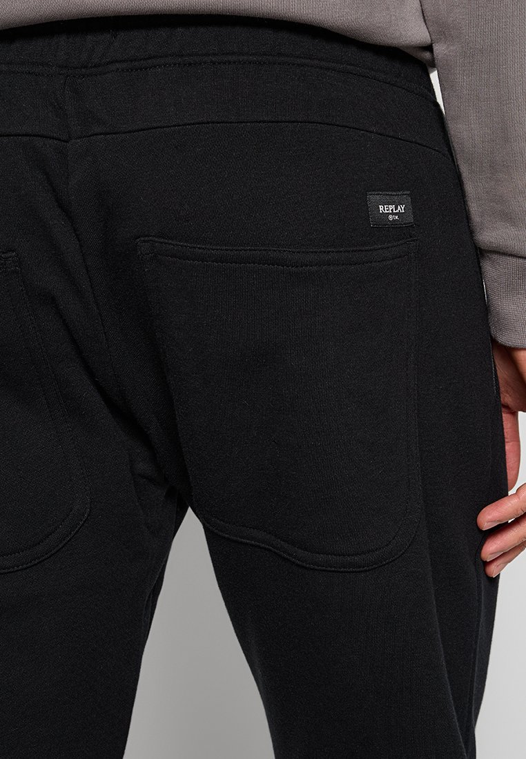 Primo piano di pantaloni neri con tasche posteriori e un piccolo etichetta del marchio "Replay" sulla tasca destra, indossati con una camicia a maniche lunghe grigia.