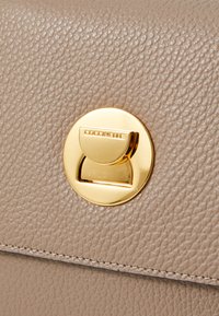 Portefeuille en cuir texturé beige avec un fermoir circulaire en or gravé "Coccinelle". Présente des coutures visibles le long des bords.