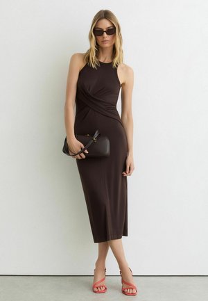 Kobieta w ciemnobrązowej, bezrękawnikowej sukience midi trzyma czarną kopertówkę, nosi czarne okulary przeciwsłoneczne i czerwone sandały na obcasie z paseczkami, stoi przy białej ścianie.