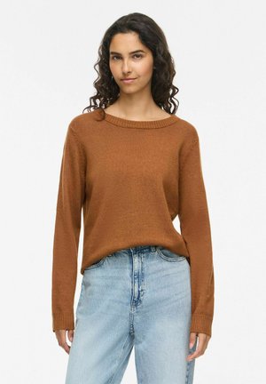 Donna con capelli scuri e ricci che indossa un maglione marrone e jeans azzurri a vita alta su uno sfondo bianco uniforme.