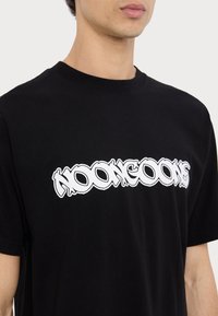 Černé bavlněné tričko s výrazným bílým grafickým nápisem "KOONGOONS" na hrudi, klasickým kulatým výstřihem a krátkými rukávy.