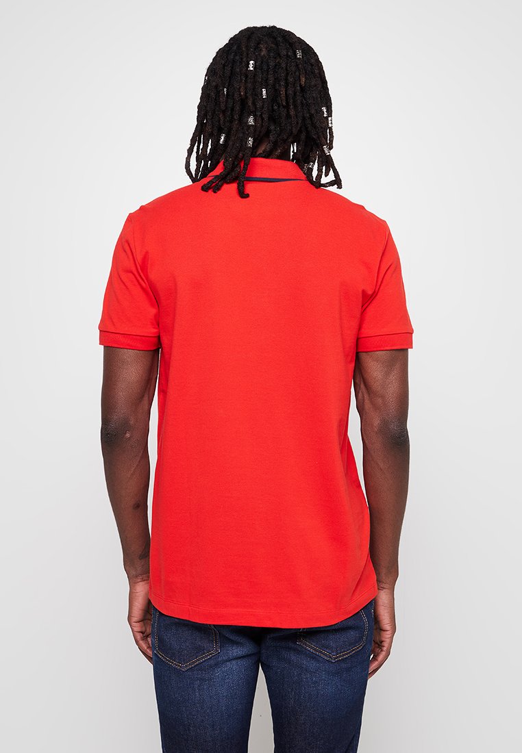 Polo rouge à manches courtes, avec col et finitions contrastées. Fait en coton, il présente une texture lisse et une coupe régulière. Porté avec un jean bleu foncé.