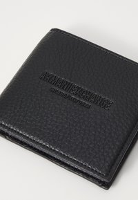 Crni kožni novčanik s teksturiranom površinom, s utisnutim brendiranjem "Armani Exchange" na prednjoj strani i preklopnim dizajnom.