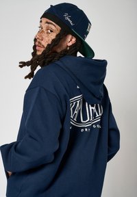 Un sweat à capuche bleu marine avec un grand graphique blanc dans le dos, affichant le mot "Kaporal". Accompagné d'une casquette assortie bleu marine, les deux fabriqués en tissu.