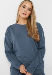 Sudadera azul con puños acanalados y cuello redondo. Textura suave, corte holgado y mangas largas, con un sutil patrón horizontal.