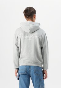 Sudadera con capucha gris de corte relajado, que presenta un gran logo blanco estampado en la parte posterior. Tejido texturizado y capucha espaciosa.