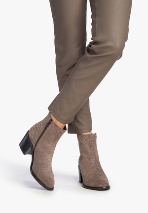 Botas marrones hasta el tobillo hechas de ante texturizado con un patrón de cocodrilo, con cierre de cremallera y un tacón grueso. Combinadas con pantalones marrones ajustados.