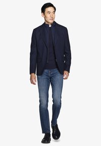 Marineblazer over en marineblå lynlåstrøje, parret med blå jeans. Modellen har sorte snørebåndsstøvler på. Glat stof, skræddersyet pasform, moderne design.