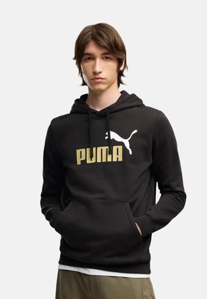 Giovane uomo con capelli castani che indossa una felpa nera Puma con logo dorato e pantaloni kaki, mani nella tasca frontale, espressione neutra.