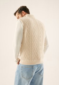 Veste en tricot beige à motif torsadé, col montant et ourlet élastique, associée à une chemise blanche à manches longues et un jean bleu clair.