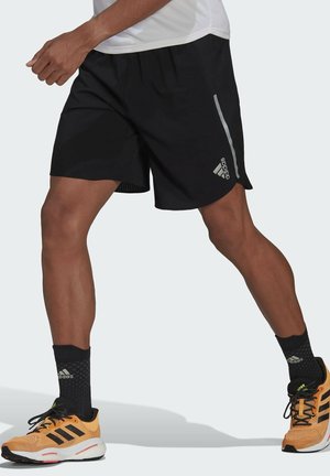 Shorts sportivi neri con vita elastica, pannello in rete laterale e striscia riflettente. Indossati con scarpe da ginnastica arancioni e calzini neri.
