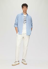 Helles blaues langärmliges Shirt über einem weißen T-Shirt mit schwarzem Text, cremefarbene Hose und beigefarbene Sneaker mit Akzenten. Glatte Stofftextur.