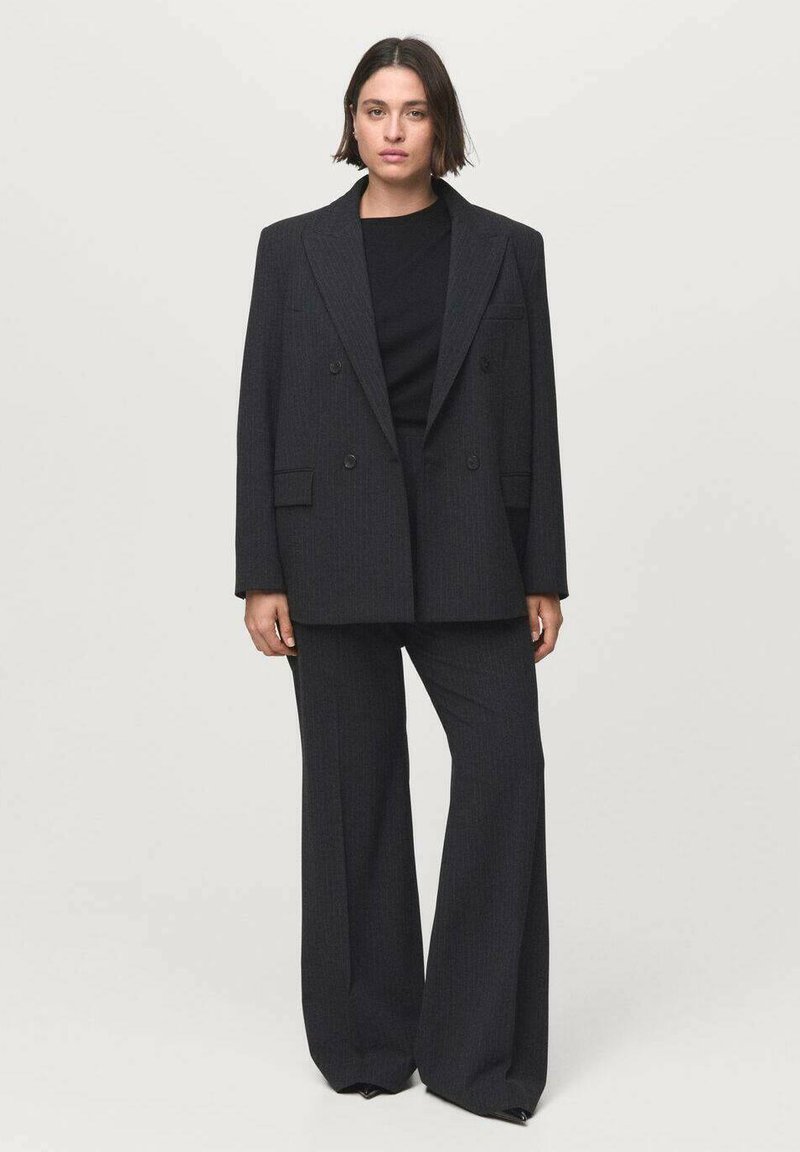 Donna che indossa un blazer oversize a righe sottili scure e pantaloni larghi abbinati su una maglietta nera, in piedi davanti a uno sfondo grigio chiaro uniforme.