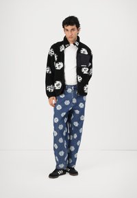 Black fuzzy jacket with white dice pattern, paired with blue jeans featuring a similar dice print. Model wears a white shirt and black sneakers.