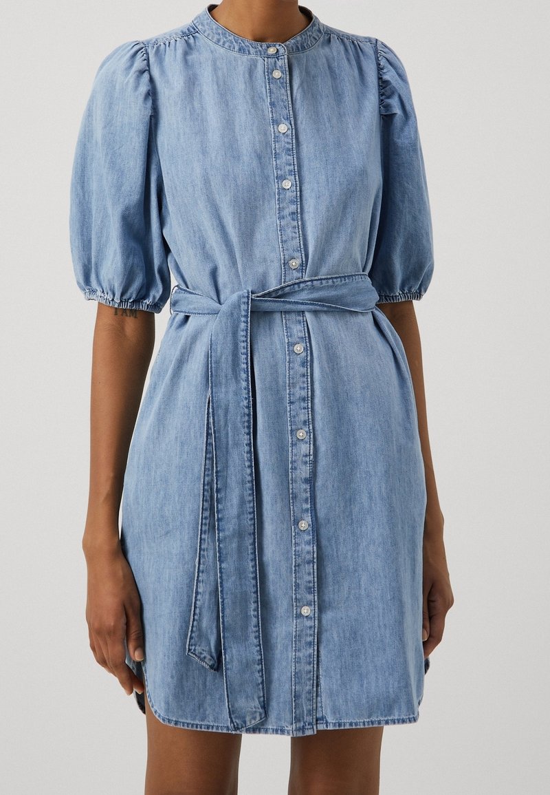 Robe en jean avec manches courtes bouffantes, boutons sur le devant, col et ceinture nouée à la taille. Couleur bleu clair avec une texture lisse.