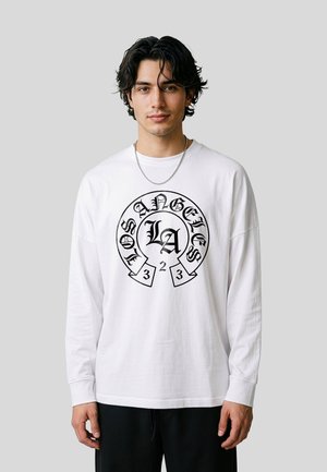 Jeune homme portant une chemise blanche à manches longues avec un motif texte noir "Los Angeles LA 2323", debout devant un fond gris clair uni.