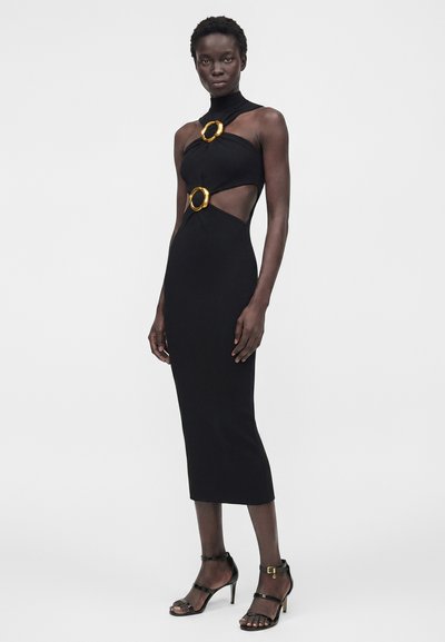 Γυναίκα που φοράει μαύρο midi αμάνικο φόρεμα με χρυσές λεπτομέρειες σε σχήμα δαχτυλιδιού στο στήθος και τη μέση, συνδυασμένο με μαύρα ψηλοτάκουνα σανδάλια με λουράκια.