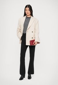 Beige, dupla soros blézer egy fekete-fehér csíkos ing felett, fekete, harang szabású nadrág és fekete magassarkú csizma; egy bordó clutch-ot tart.
