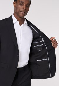 Schwarze Sakkogjacke aus glattem Stoff, mit einem weißen Hemd, Knopfverschluss und Innentaschen mit hellgrauen Akzenten.