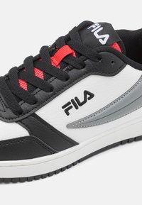 Zapatilla en blanco y negro con cuero texturizado, acentos plateados y lazos de encaje rojos. Presenta una lengua acolchada y un logo en el lateral.