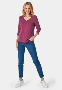 Pflaumen-farbenes V-Ausschnitt-Oberteil mit drei Viertelärmeln, kombiniert mit blauen Skinny-Jeans und weißen Sneakers. Glattes Material und taillierte Passform.