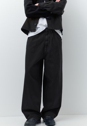 Person iført løse sorte jeans, sort jakke, hvid skjorte delvist ude af bukserne, stående med armene krydsede mod en ensartet hvid baggrund.