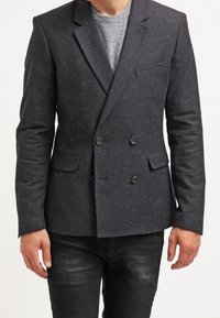 Granatowa marynarka dwurzędowa z subtelnymi białymi kropkami, z wyciętymi klapami i bocznymi kieszeniami, w połączeniu z jasnoszarym swetrem i ciemnymi dżinsami.