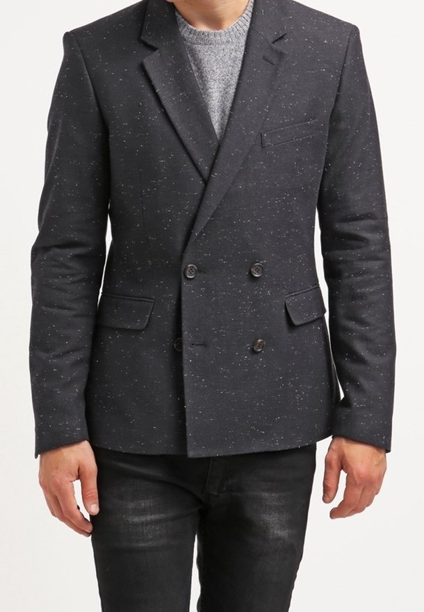 Granatowa marynarka dwurzędowa z subtelnymi białymi kropkami, z wyciętymi klapami i bocznymi kieszeniami, w połączeniu z jasnoszarym swetrem i ciemnymi dżinsami.