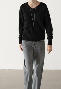 Pull noir en v en matière douce, avec des manches longues. Assorti à un jean droit gris clair et un long collier en pendentif argenté.