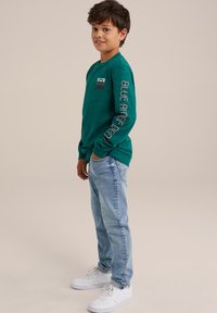 Långärmad turkos skjorta med en liten logotyp på bröstet och text på ärmen, matchad med ljusblå jeans och vita sneakers.