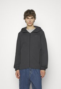 Dunkelgraue Kapuzen-Zip-Jacke mit glatter Textur, ausgestattet mit Seitentaschen und dezenten Nähten, kombiniert mit blauen Jeans.