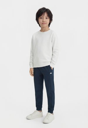Child standing with hands in pockets, wearing white sweatshirt, navy blue joggers, and white sneakers on a plain white background.