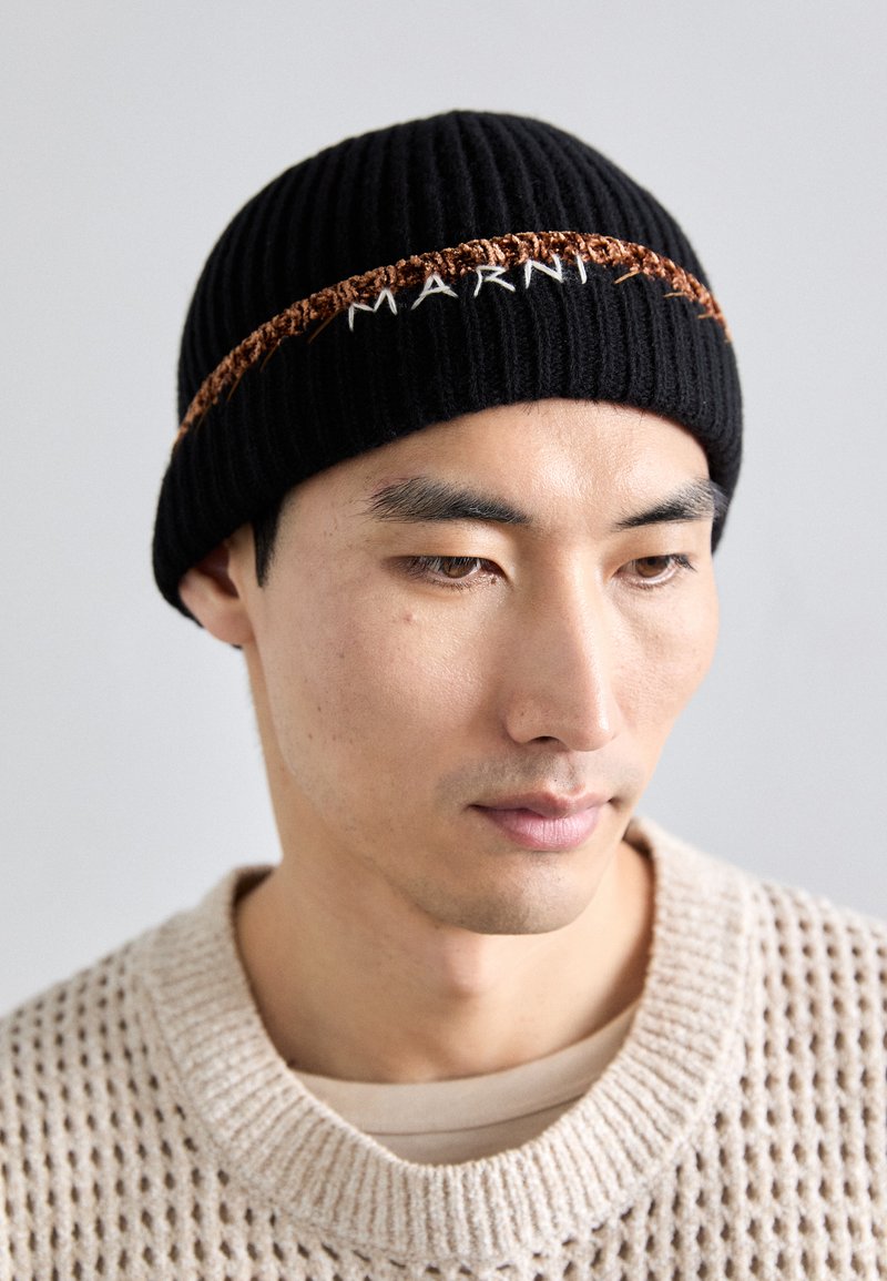 Ung mand iført en sort ribbenstrik-hat med "MARNI" broderi og en beige tekstureret sweater, kigger ned mod en ensfarvet baggrund.