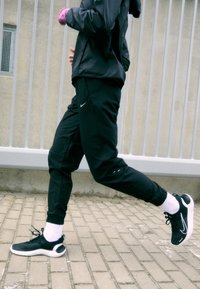 Czarna sportowa kurtka i spodnie, czarne sneakersy z białymi podeszwami i białe skarpetki. Biegacz biegający wzdłuż ceglanej ścieżki, o opływowym kształcie.