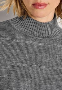 Frau trägt einen grauen, gerippten Strickpullover mit Rollkragen, der den Hals, das Untergesicht und die Strukturdetails des Stoffes zeigt.