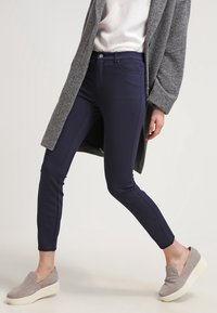 Marina blå slim-fit byxor tillverkade av stretchigt tyg, med en mjuk yta och en klassisk femficksdesign, tillsammans med grå slip-on skor.