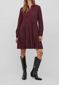 Abito button-up bordeaux con maniche lunghe e gonna a strati. Tessuto testurizzato, lunghezza al ginocchio, abbinato a stivali neri in pelle alti.