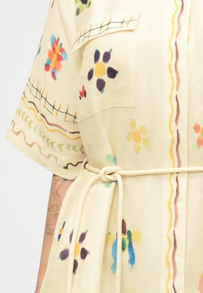 Φωτεινό μπεζ φόρεμα με πολύχρωμα λουλουδένια μοτίβα, κοντά μανίκια, μπροστινή τσέπη και λεπτή ζώνη δεμένη στη μέση, φορεμένο από έναν τατουαρισμένο βραχίονα.
