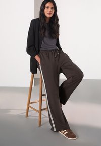 Braune Sweatpants mit weitem Bein und weißen Seitenstreifen, kombiniert mit einem grauen Oberteil und einem schwarzen Blazer. Dazu braune Sneakers. Auf einem Holzstuhl sitzend.