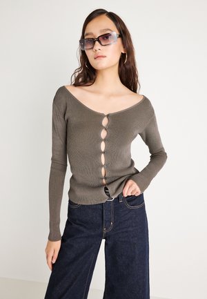 Young woman wearing oversized rectangular sunglasses, olive ribbed buttoned sweater with cutouts, and dark wide-leg jeans against plain background.